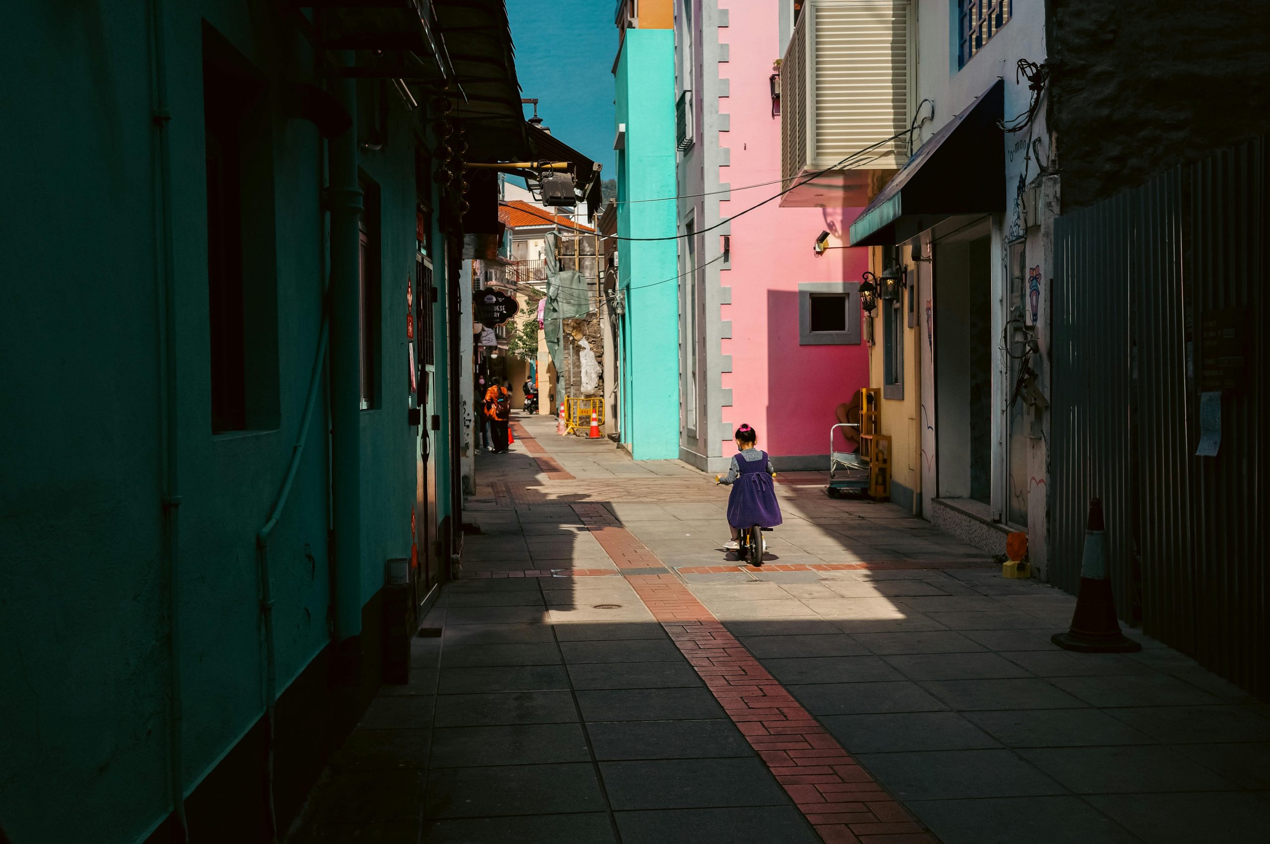 A child on a scooter explores a vibrant alleyway in Macau, filled with colorful buildings and warm sunlight.