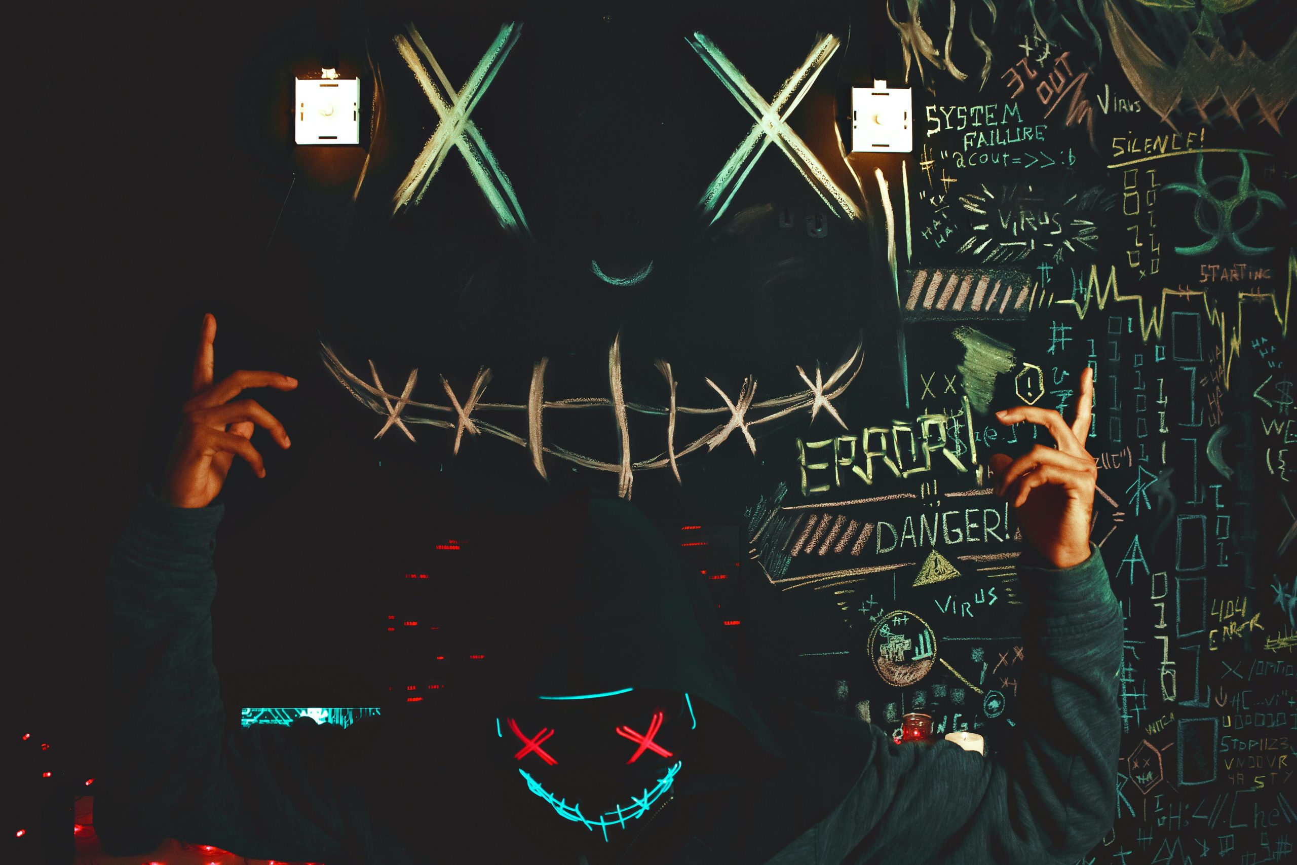Intriguing hooded individual with neon mask against a chalkboard backdrop of cryptic symbols.