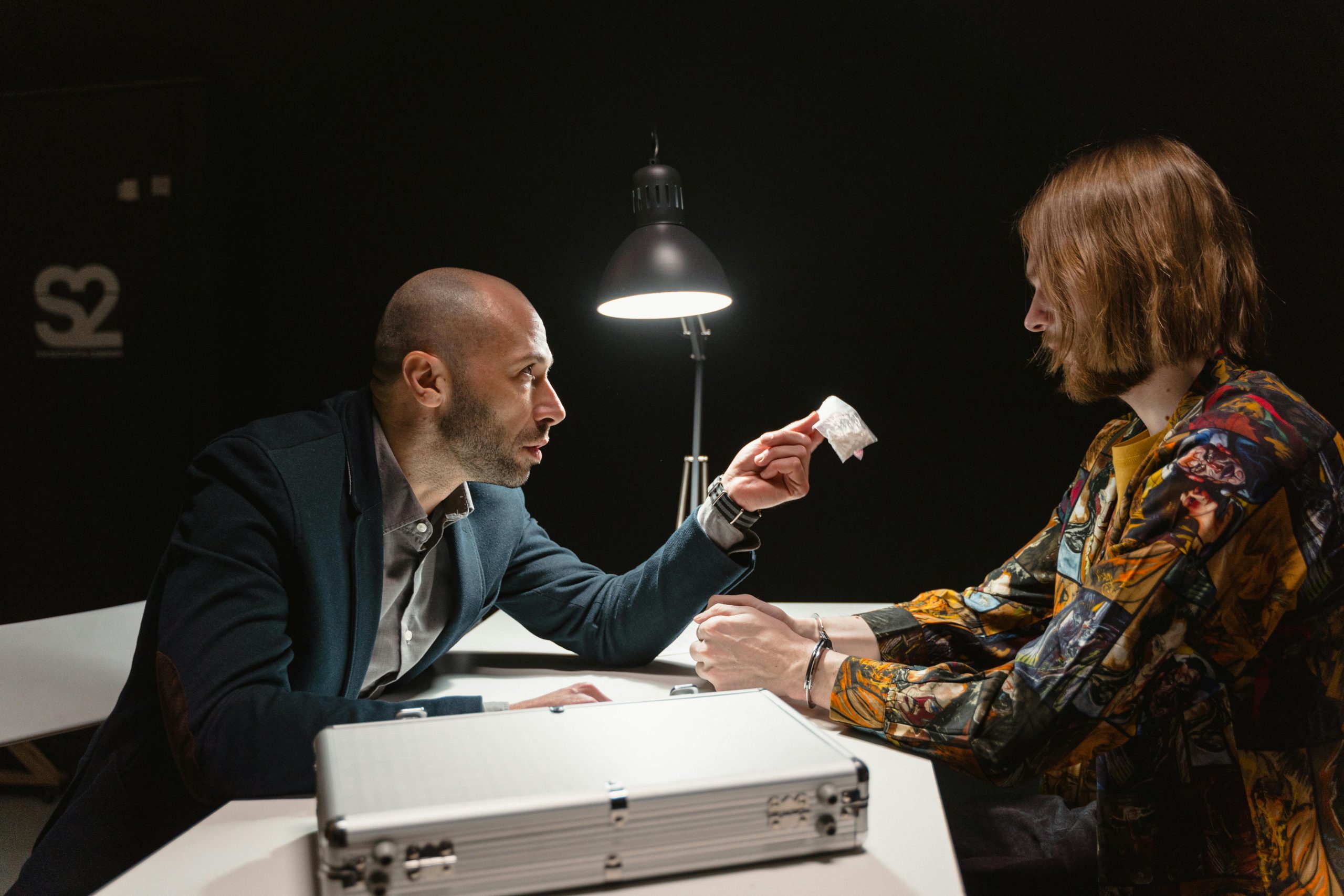 Two adults in a dimly lit room negotiating over a small package at a table.